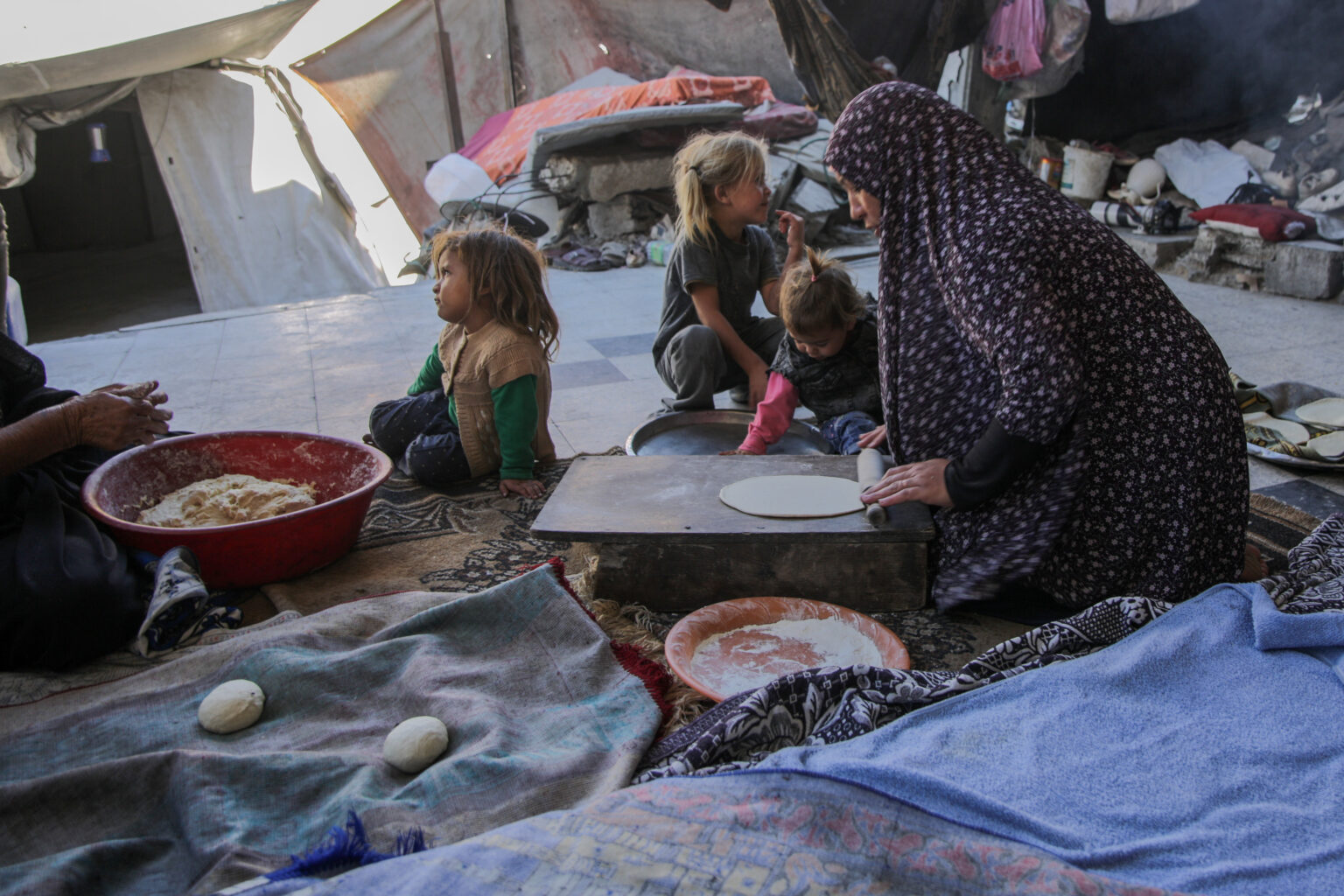 Una donna seduta per terra fra le macerie vicino la sua tenda cerca di fare il pane, accanto a lei ci sono tre bambini piccoli.