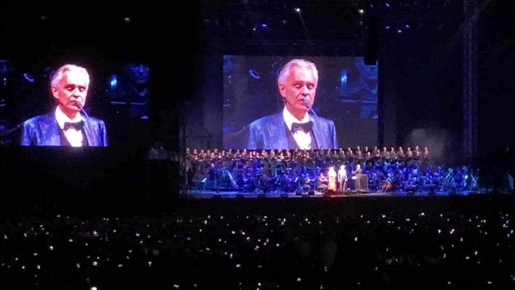 Andrea Bocelli a mezzo busto e la sua immagine riflessa su schermo durante un concerto a Tel Aviv.